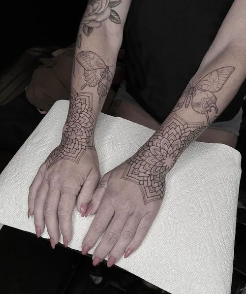 Two tattooed forearms resting on a white cushion, hands together with long manicured nails.