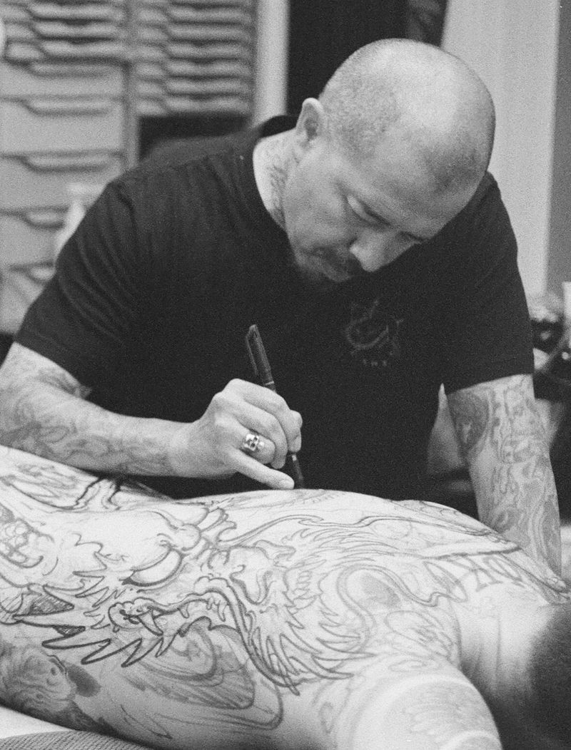 Tattoo artist sketching on a client’s back with a pen in a black-and-white close-up.