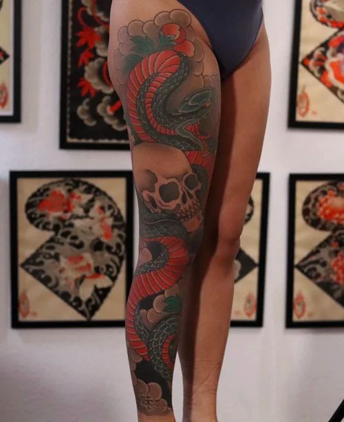 Side view of a leg tattoo featuring red and green snakes wrapping around a skull.