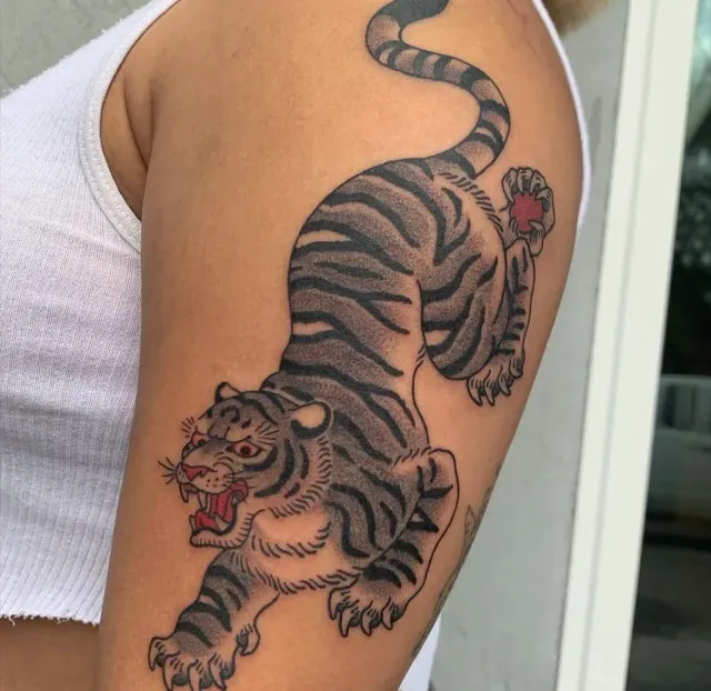 Arm tattoo of a black-and-gray tiger mid-pounce with red accents, on an upper arm.