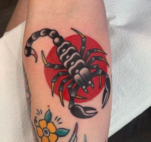 Black scorpion tattoo over a red circle on skin, with a small colorful flower tattoo below.