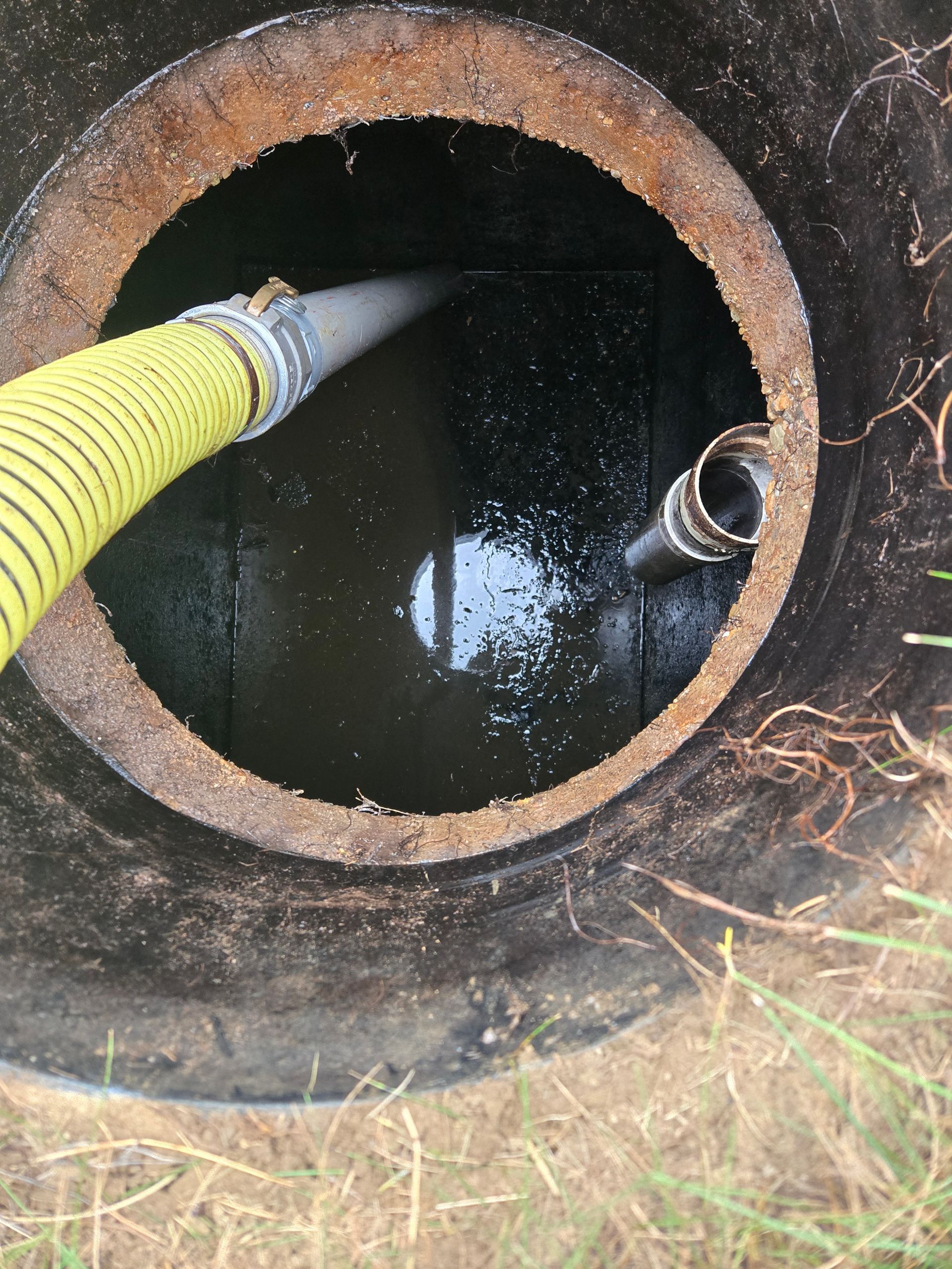 A yellow hose enters a rusty circular tank opening with dark water inside.