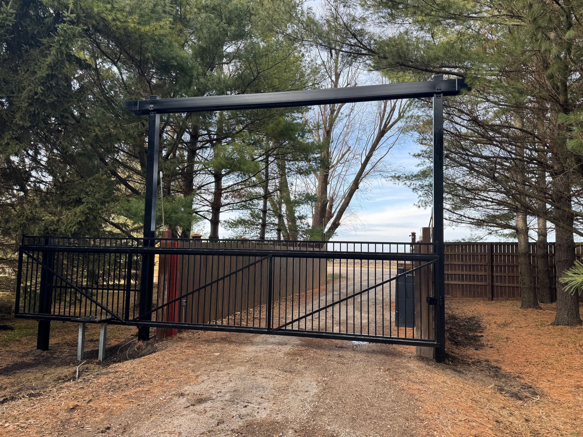 A black gate is open to a dirt road surrounded by trees.