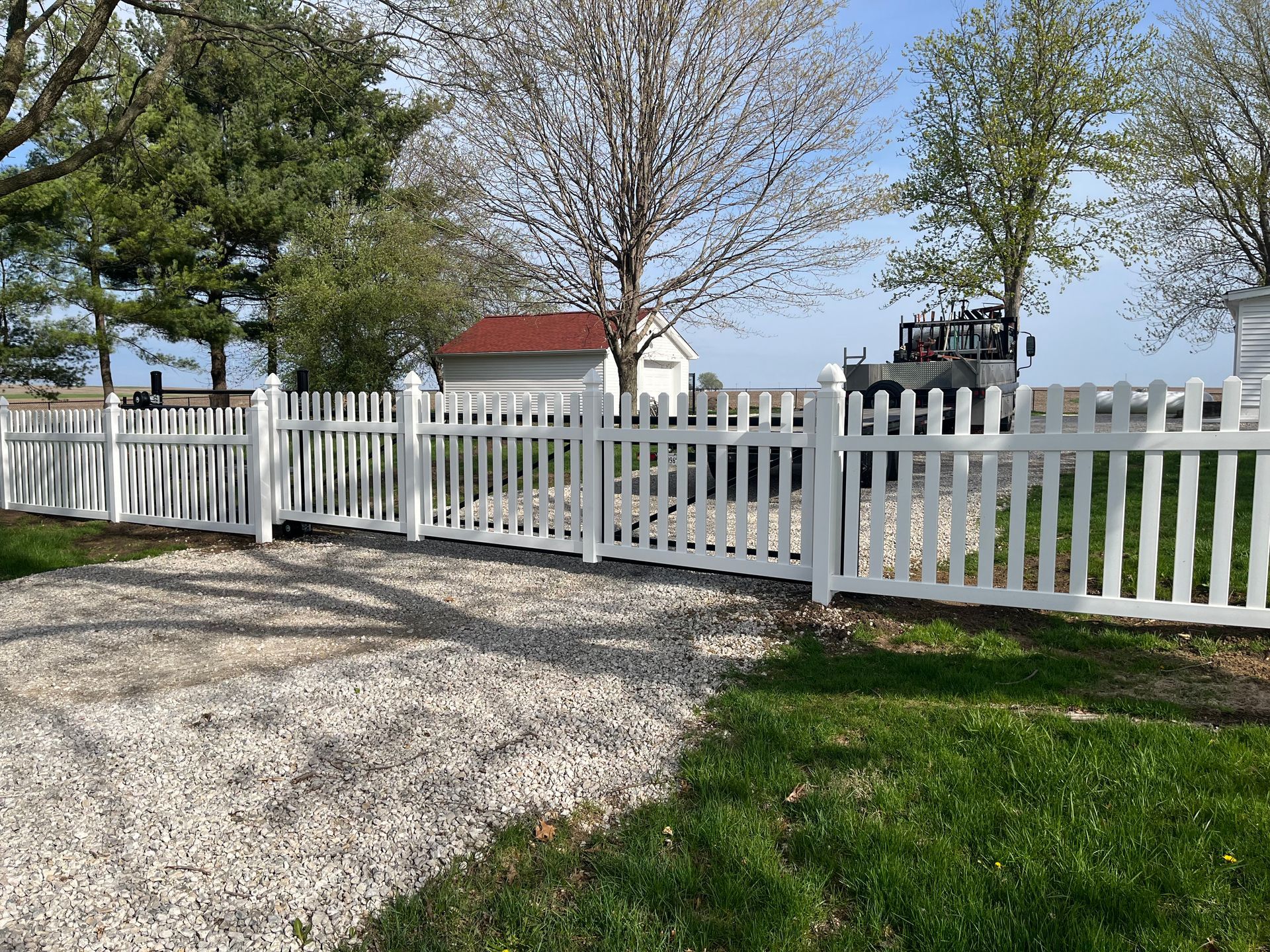 A white picket fence surrounds a gravel driveway leading to a house.
