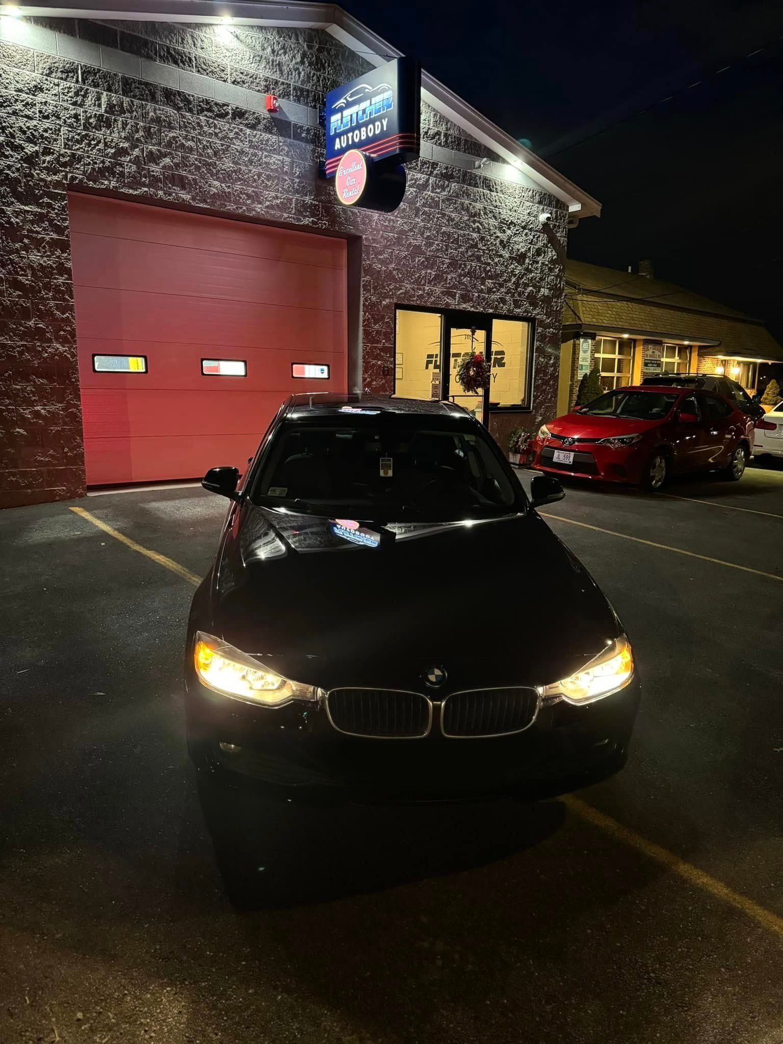 Black car parked outside a lit brick building at night, with a red garage door and illuminated sign.