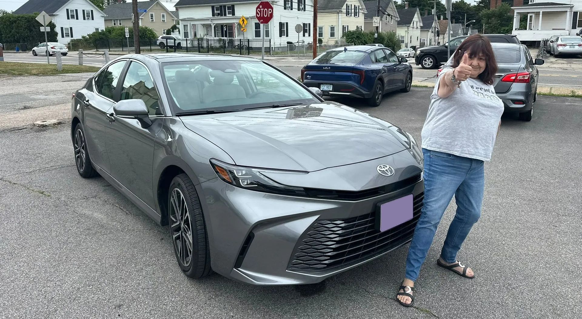 Woman posing beside a gray sedan in a parking lot, holding up a peace sign.