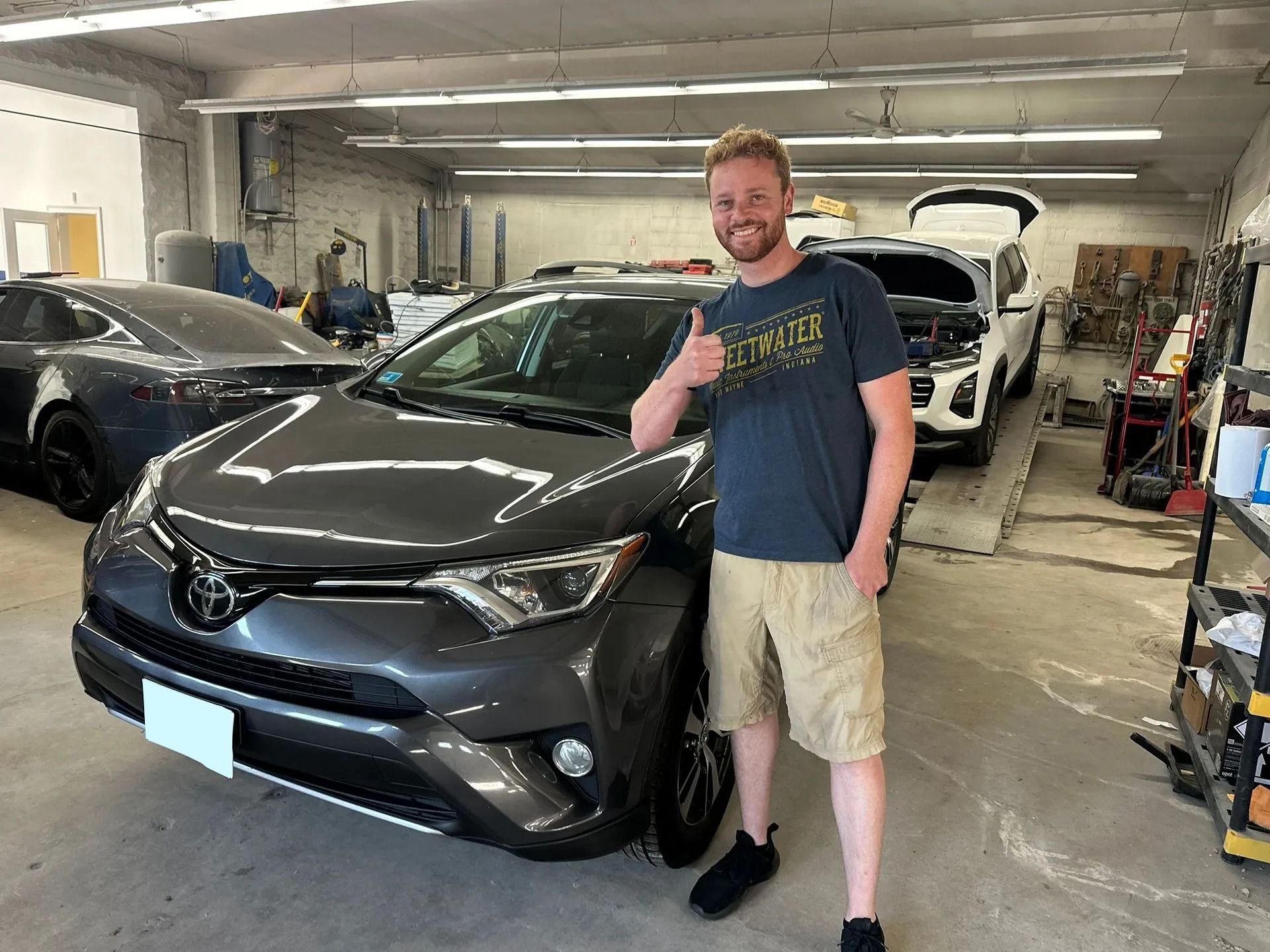 Man standing beside a gray car in a garage, giving a thumbs-up.