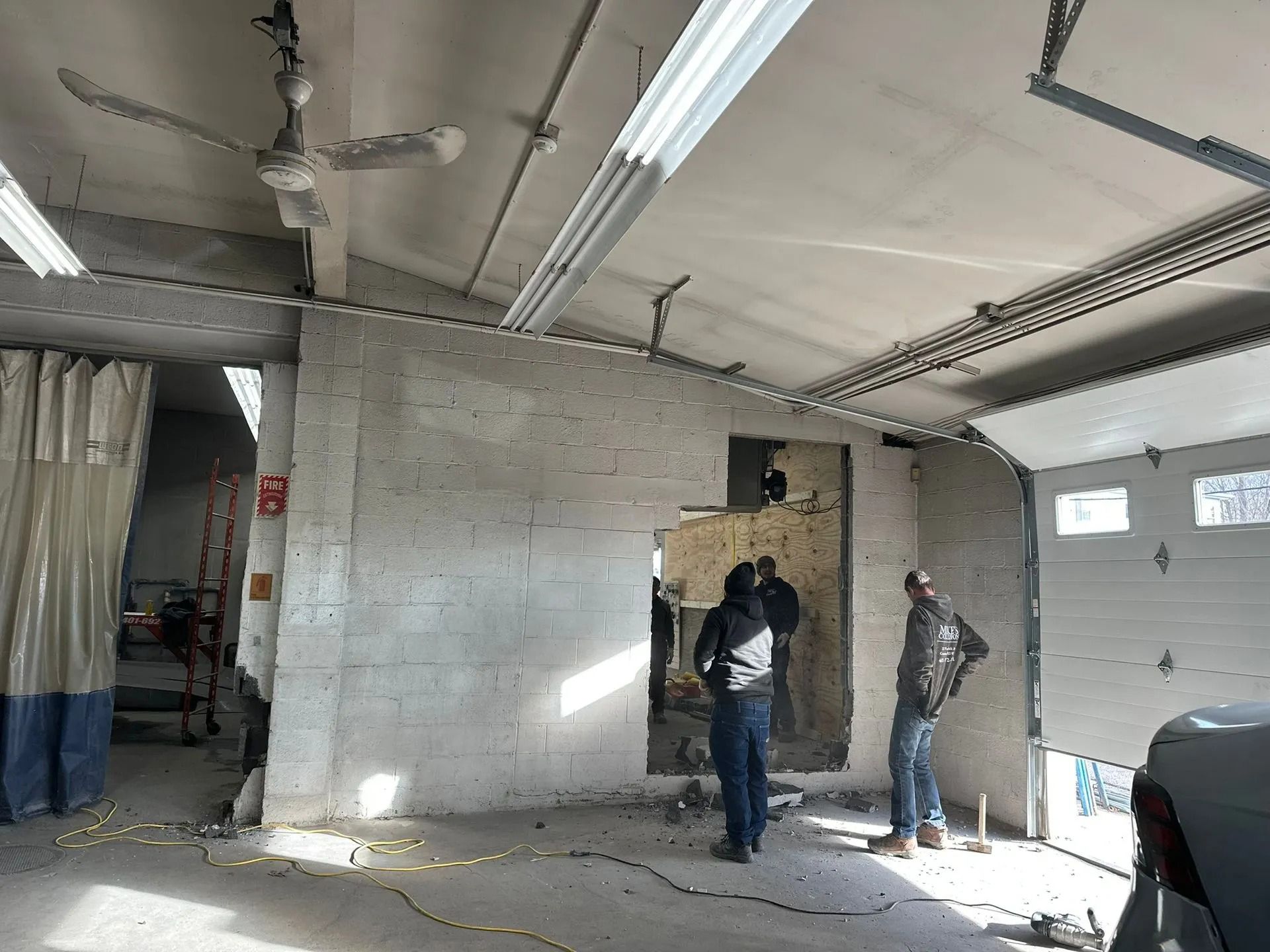 Workers installing drywall in an unfinished room with exposed ceiling beams and hanging lights.