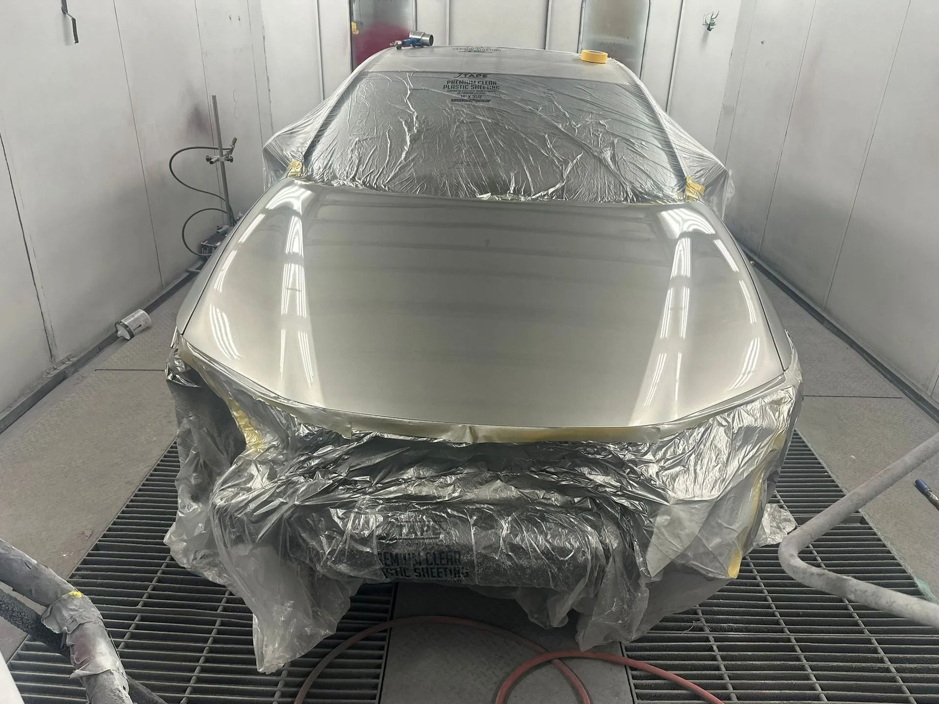 Silver car wrapped in plastic inside a spray booth, with front bumper removed