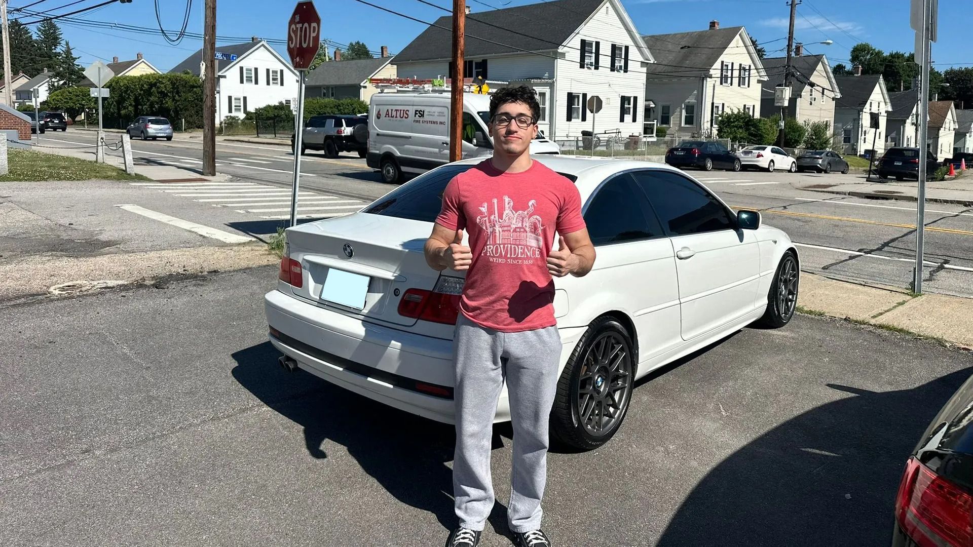 Person standing beside a white car on a sunny residential street.