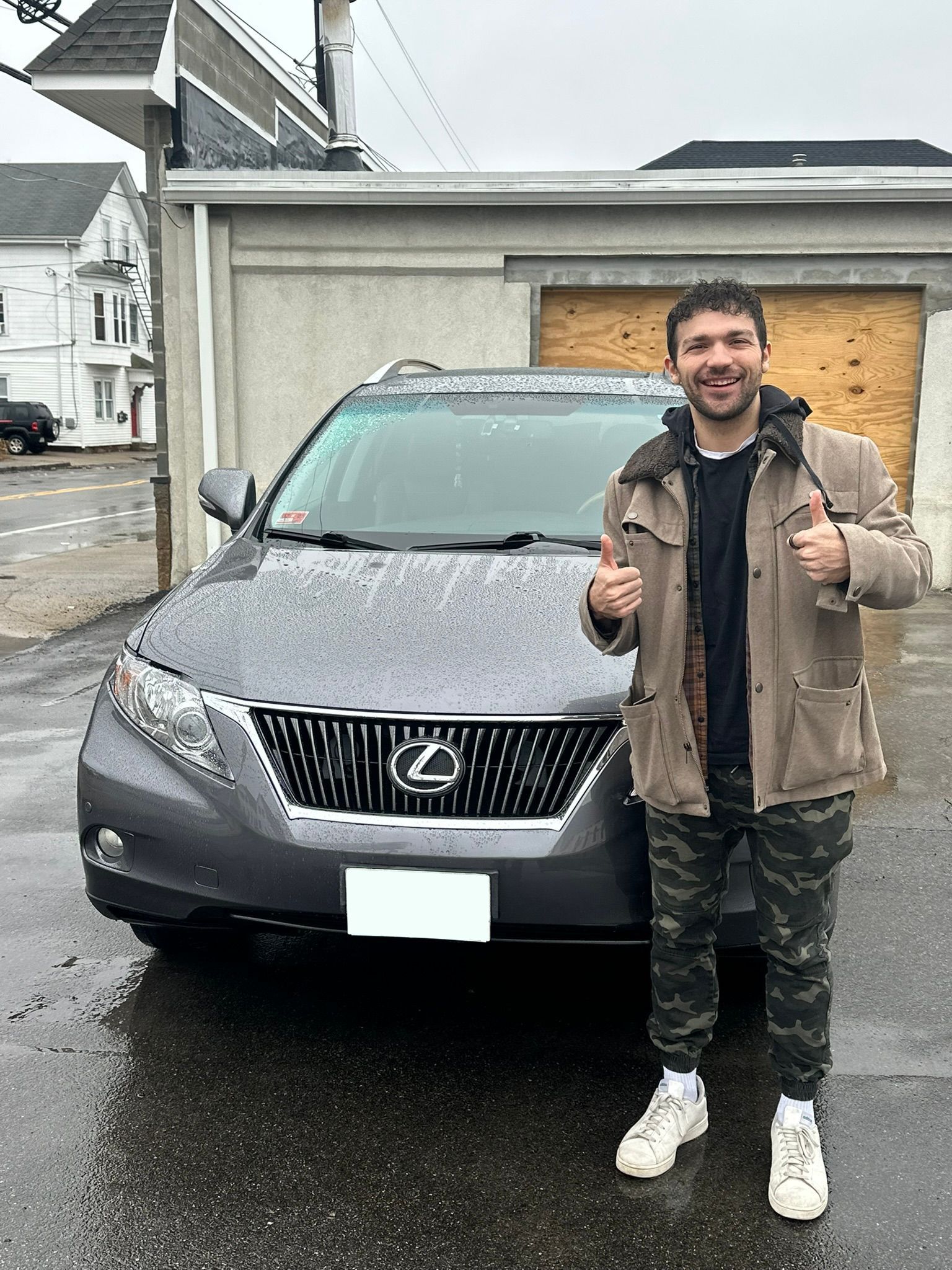 Man standing beside a gray Lexus SUV in front of a house, smiling and giving thumbs up on a rainy day