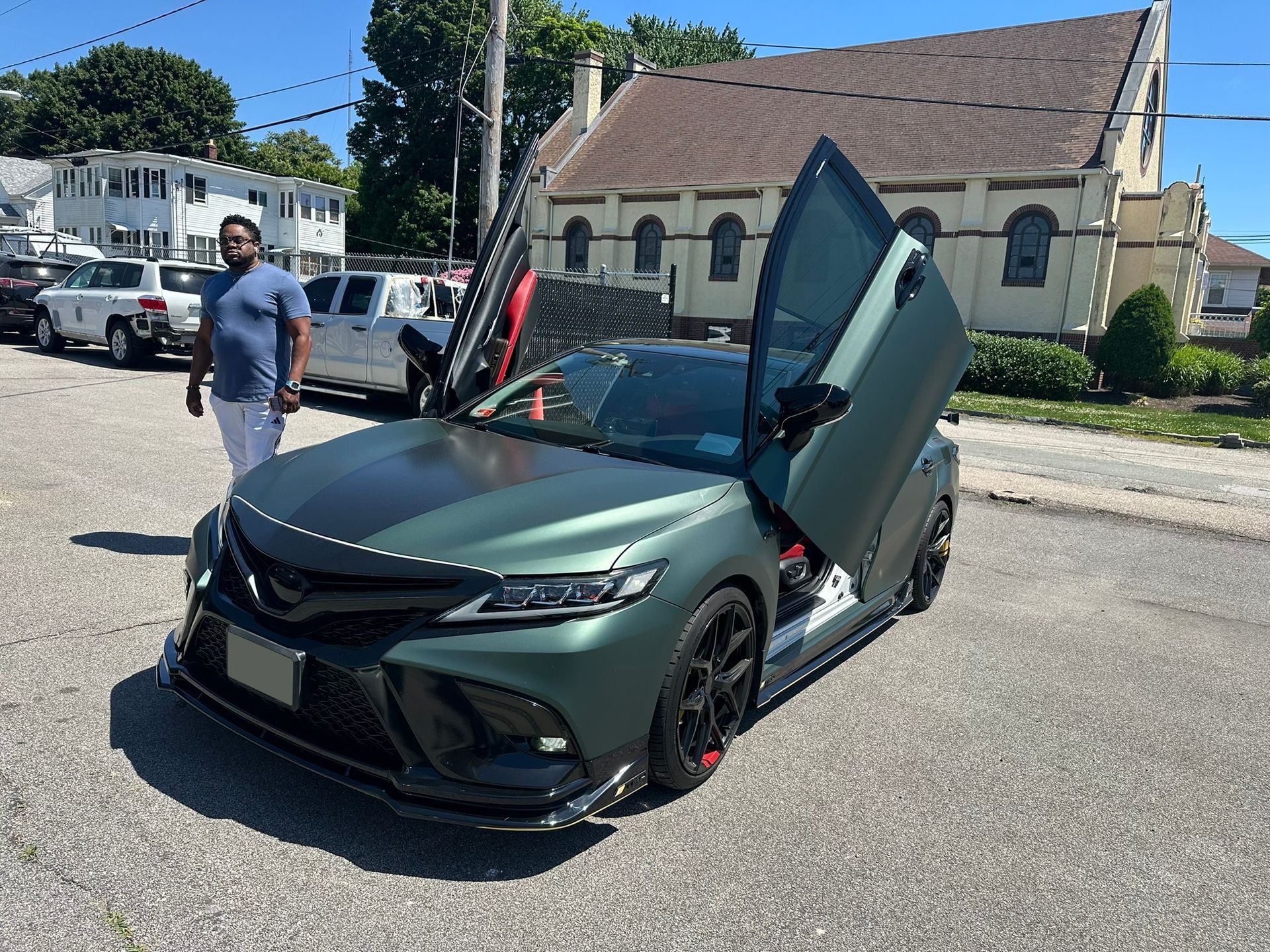 Matt green sports car with scissor door open on a street beside a man, near a church-like building