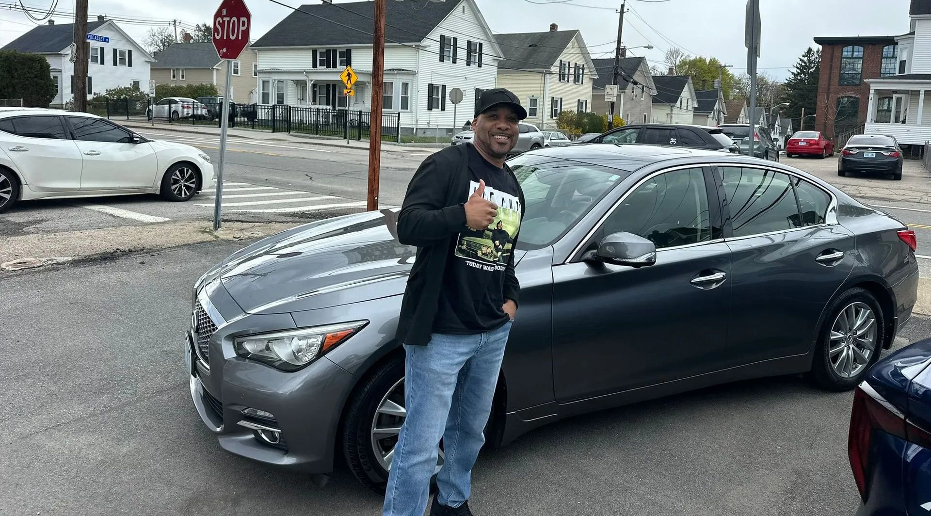 Man standing beside a gray sedan on a residential street, with houses and parked cars in the background.
