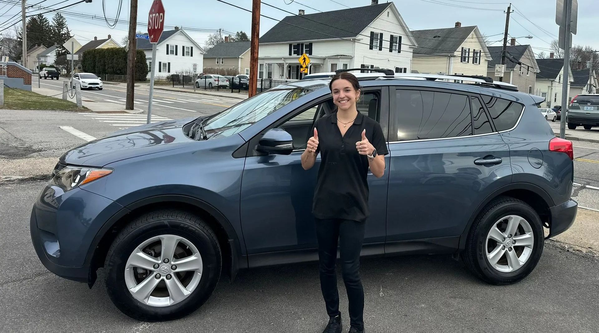 Person standing beside a blue SUV in a parking lot, giving a thumbs-up.