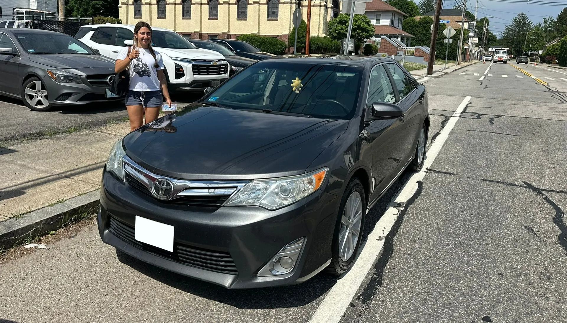 Gray sedan parked on a sunlit street with a woman standing beside it near other parked cars.