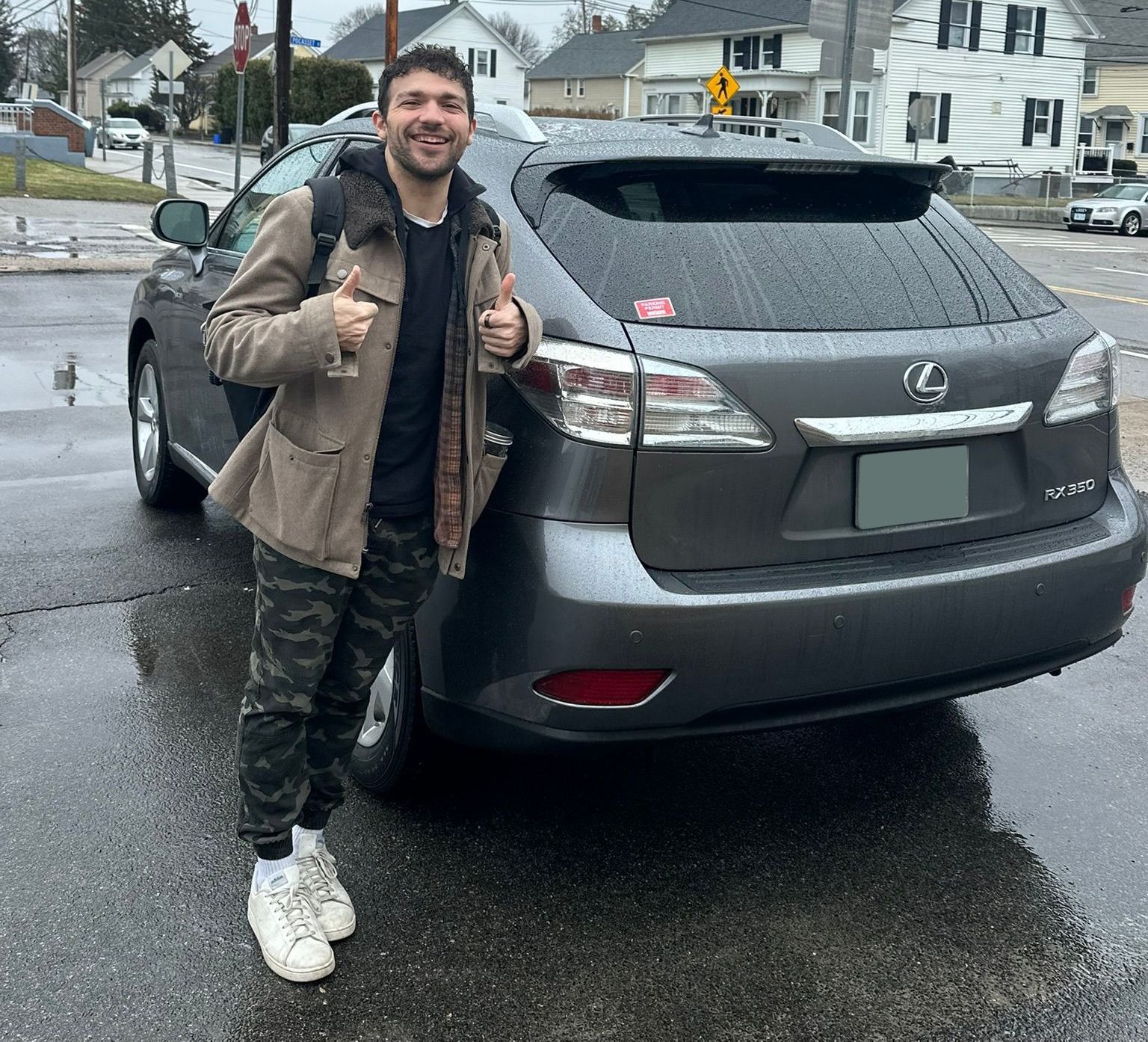 Man standing beside a gray Lexus SUV on a wet residential street, smiling and giving thumbs up