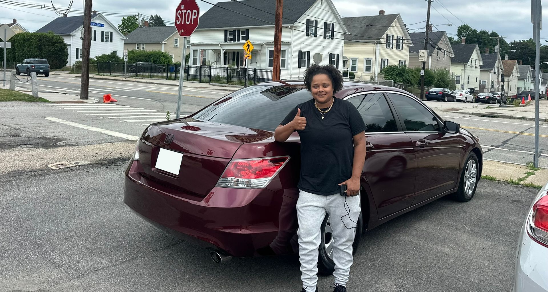 Person standing beside a maroon sedan on a residential street, with a stop sign and houses in the background.