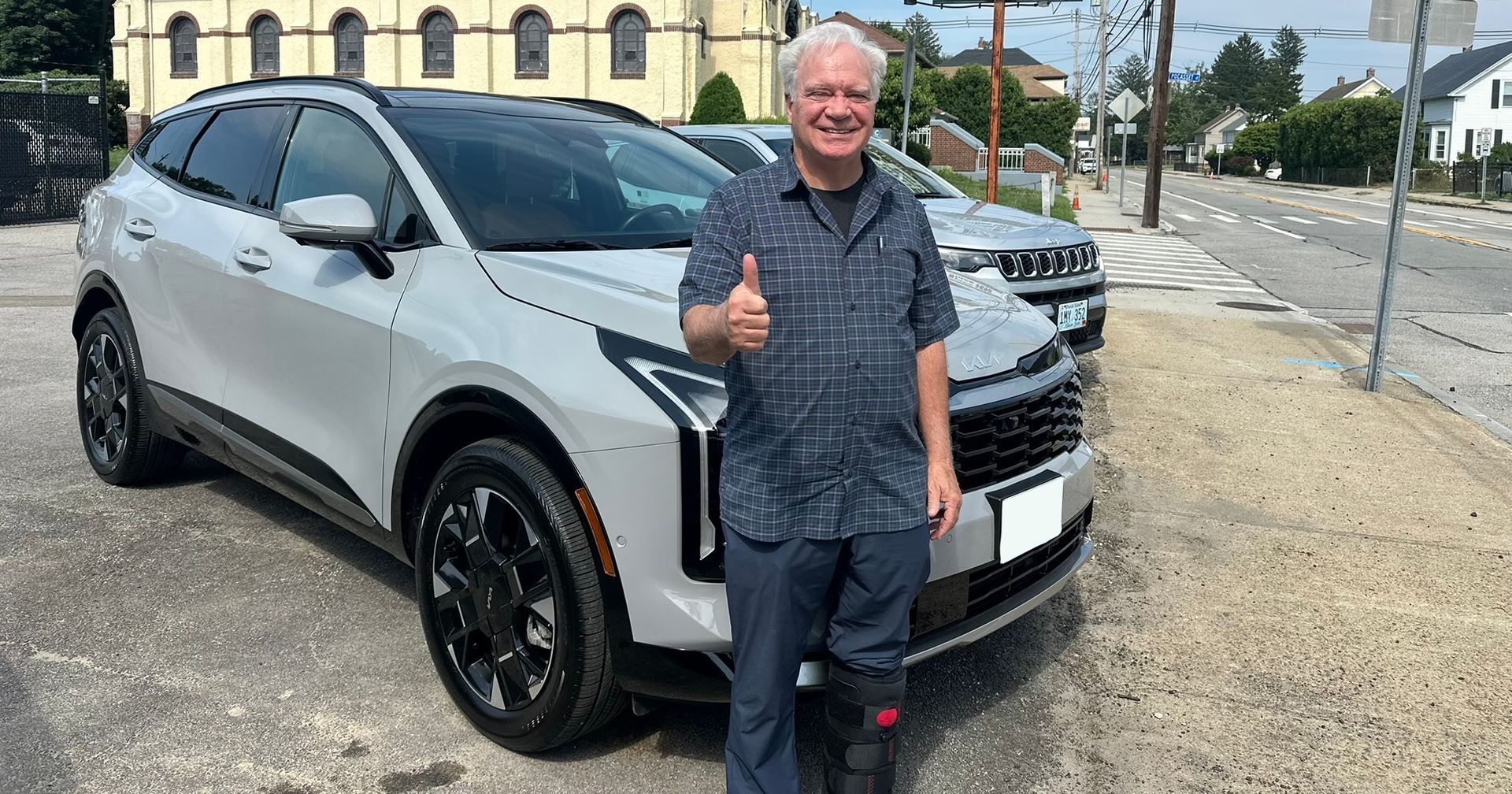Man standing beside a white SUV, smiling and giving a thumbs-up in a parking lot.