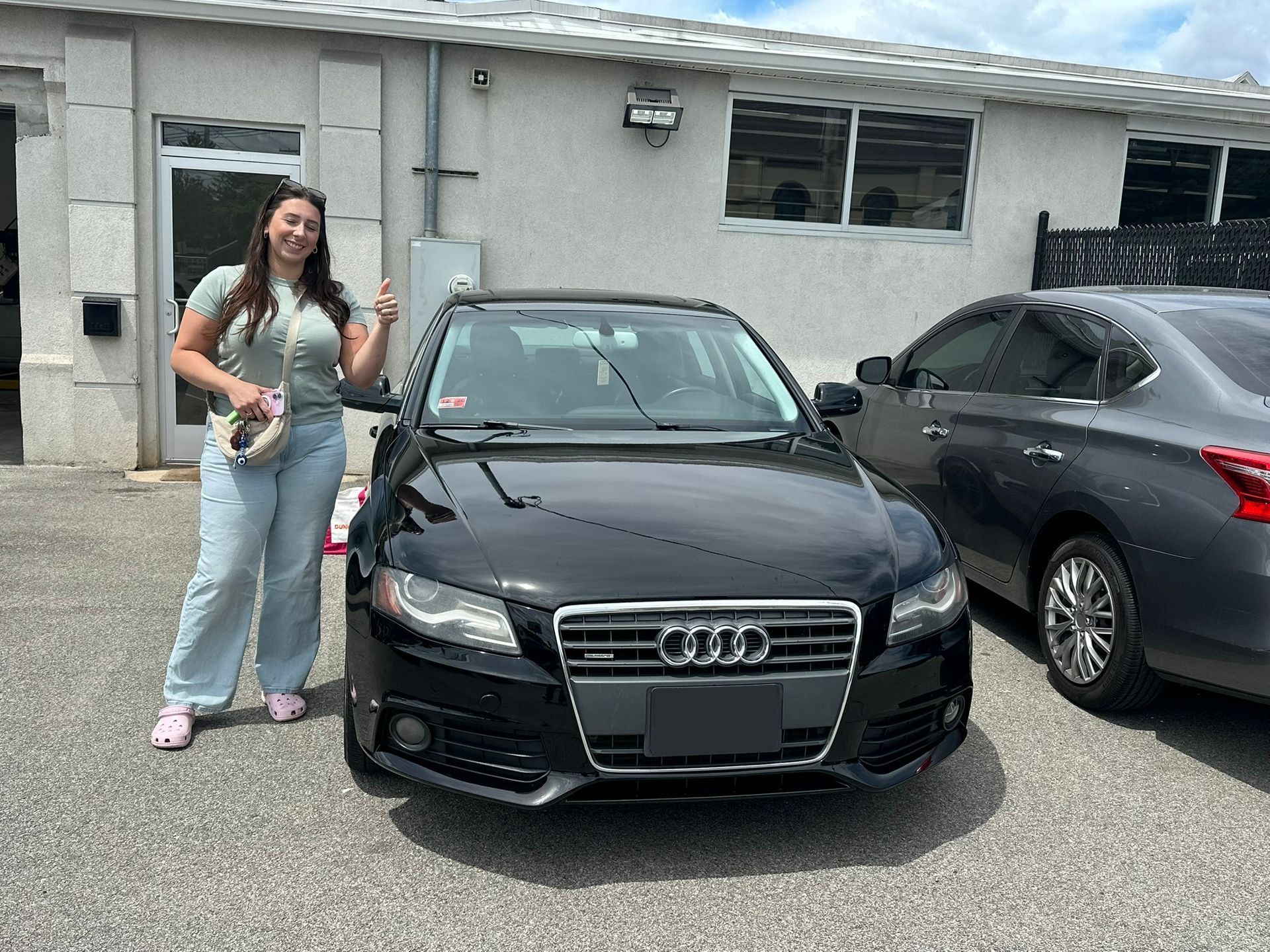 Woman standing beside a black Audi in a driveway in front of a house.
