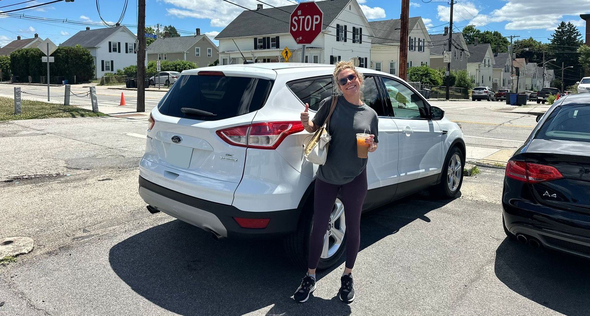 Person standing beside a white SUV at an intersection, holding a drink near a stop sign.