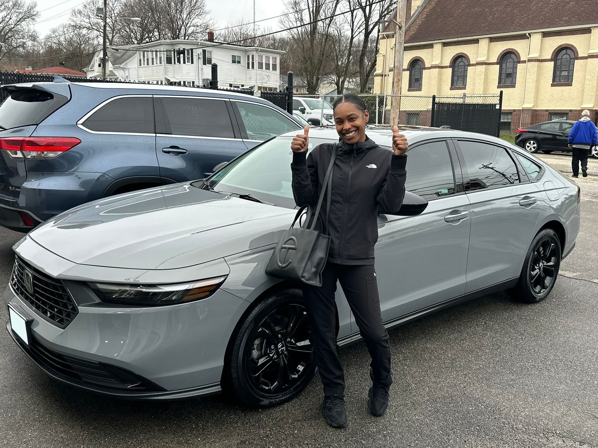 Person posing beside a gray sedan in a parking lot, smiling and holding up keys.