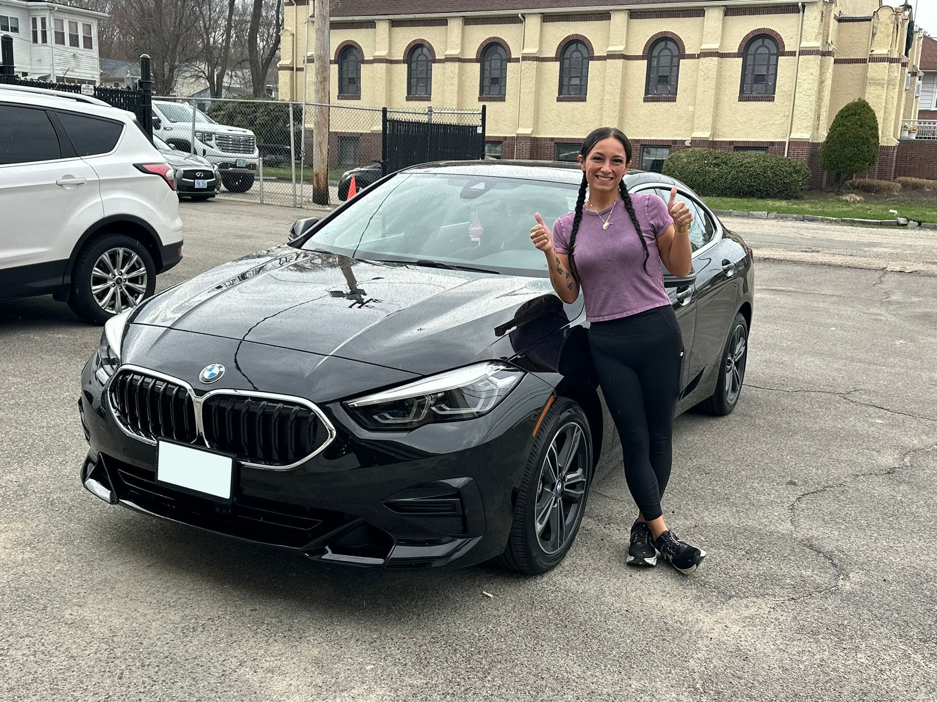 Woman leans against a black BMW in a parking lot, smiling and holding up a peace sign.