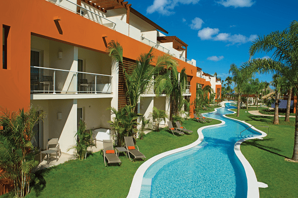 Resort buildings with balconies line a winding, blue pool. Palm trees and green grass.