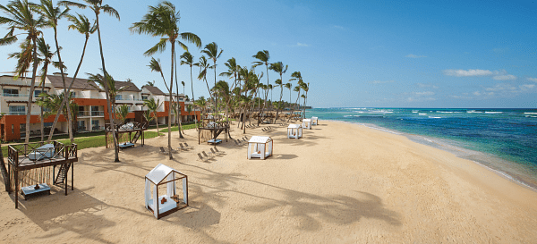 Sandy beach with palm trees, cabanas, and resort buildings under a clear blue sky.