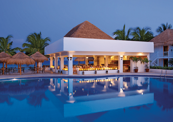 Poolside bar with thatched roof, reflecting in a blue pool at dusk, palm trees in the background.