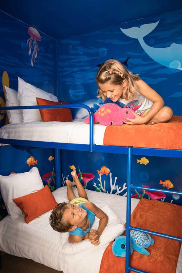 Children in bunk beds in a room with ocean-themed wall art, one child holding a toy, and the other laying down.