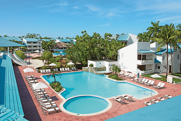 Swimming pool at resort, surrounded by white buildings, palm trees, and lounge chairs under blue sky.