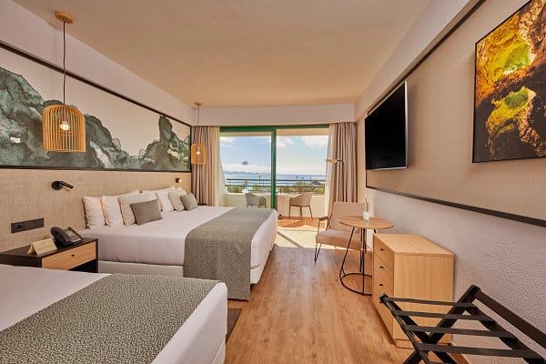 Hotel room with multiple beds, balcony view, neutral tones, wood accents.