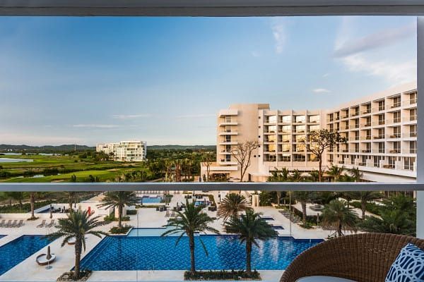 View from a balcony overlooking a resort with pools, palm trees, and a white building under a blue sky.