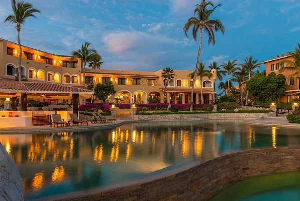 Luxury resort at dusk, buildings with warm lights reflecting in the pool with palm trees.