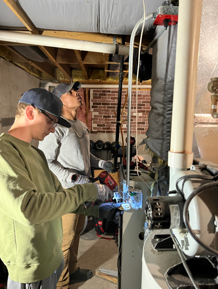 Two workers weld piping in a basement utility area, with blue sparks and exposed beams.