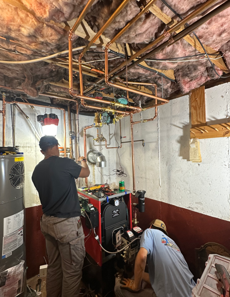 Two workers installing HVAC equipment in a small room with exposed ceiling ducts and insulation.