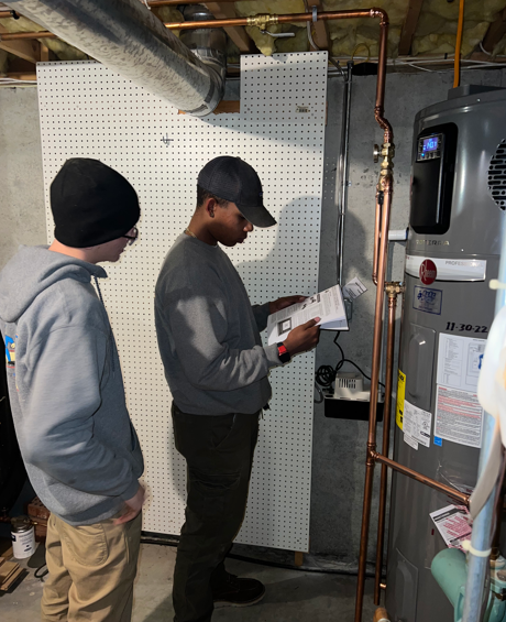 Two workers inspect a furnace and HVAC system in a basement utility room.