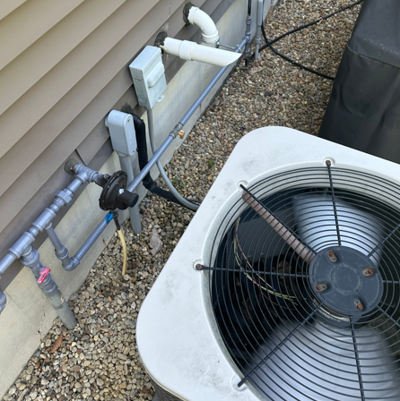 Outdoor HVAC unit beside house siding, with exposed pipes and electrical boxes on a gravel bed.