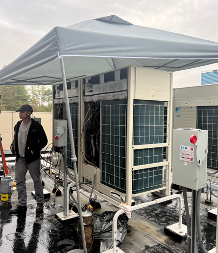 Outdoor HVAC unit under a white canopy on a wet rooftop, with a worker standing nearby.