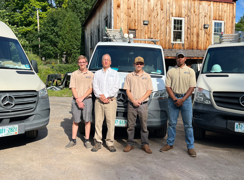 Four people standing in front of parked vans beside a wooden building outdoors