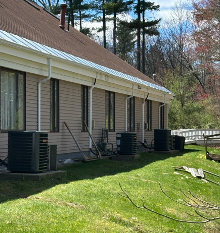 Backyard view of a beige house with brown roof, air conditioners, and a green lawn under sunny skies