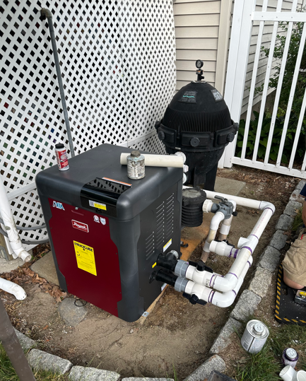 Outdoor pool equipment: red heater, black filter tank, and white PVC pipes beside a lattice fence.