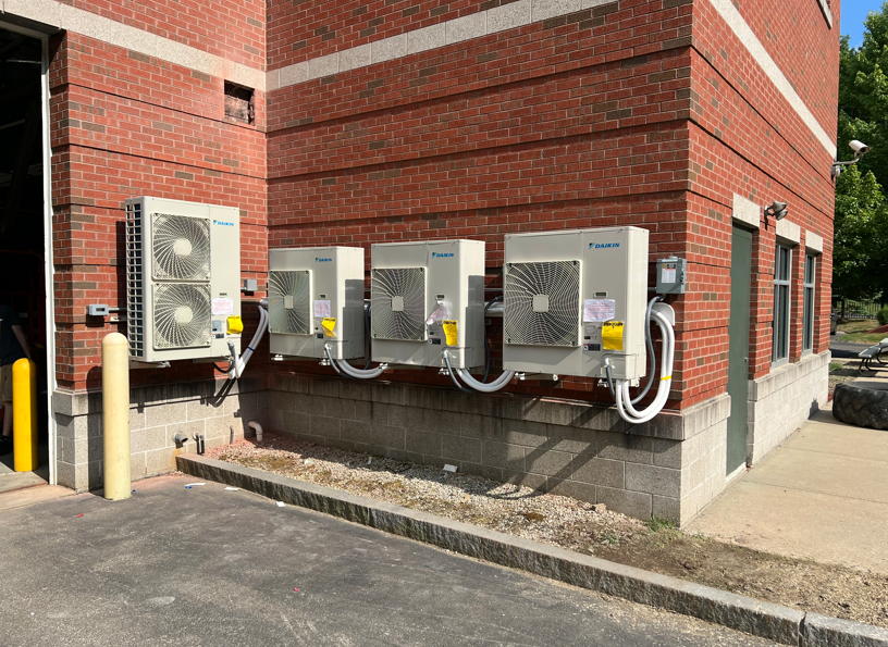 Row of outdoor HVAC units beside a red brick building, with white conduit and a gravel base.