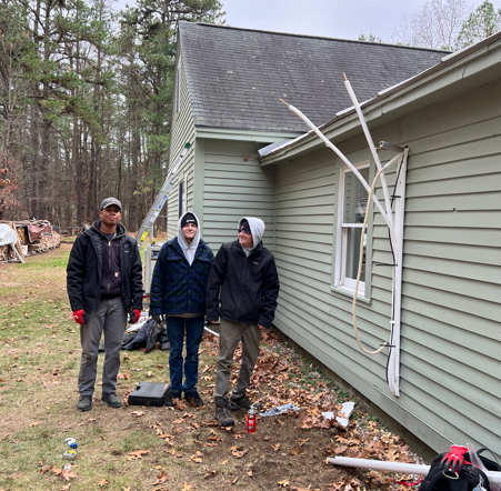 Three people stand beside a house with damaged siding and branches leaning against the wall.