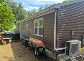 Brown house exterior with white-trim windows, AC unit, canoe, and yard equipment beside a wooded driveway