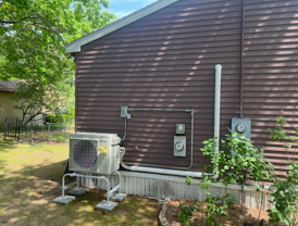 Outdoor HVAC unit beside a brown house with utility meters and a small garden bed