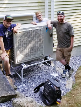 Three men posing beside an outdoor HVAC unit on gravel by a house.
