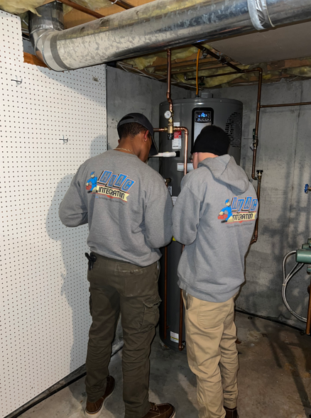 Two technicians in gray sweatshirts inspect equipment in a basement utility room.