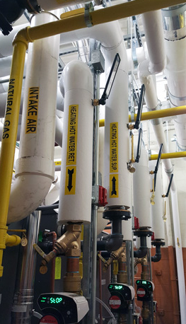 Industrial pipes and ducts with yellow labels in a mechanical room; pumps and gauges below.