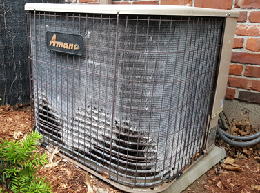Damaged Amana outdoor air conditioner unit with bent metal grille beside a brick wall and mulch bed